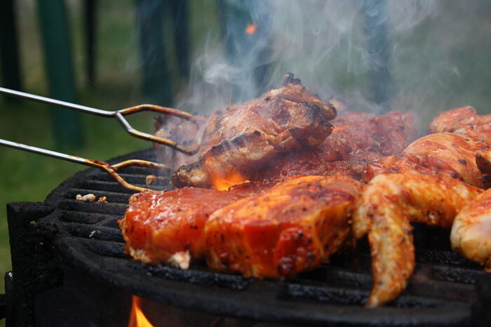 Several pieces of seasoned meat are grilled over an open flame at a private schools on the Northshore event, tongs lifting one as smoke rises.
