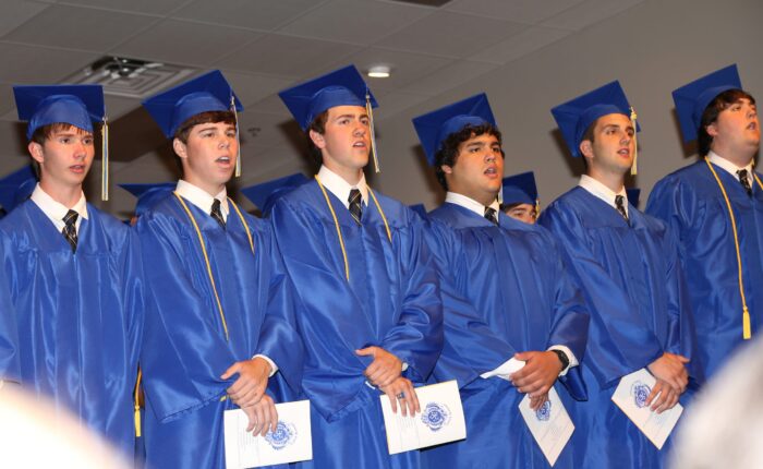 Graduates in blue caps and gowns from St. Paul's School stand in a row, holding programs during an indoor ceremony.