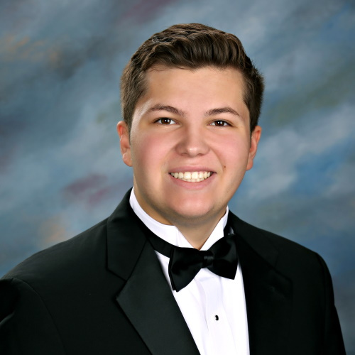 A young man in a black tuxedo and bow tie, smiling in a formal studio portrait at St. Paul's School with a blue and gray background.