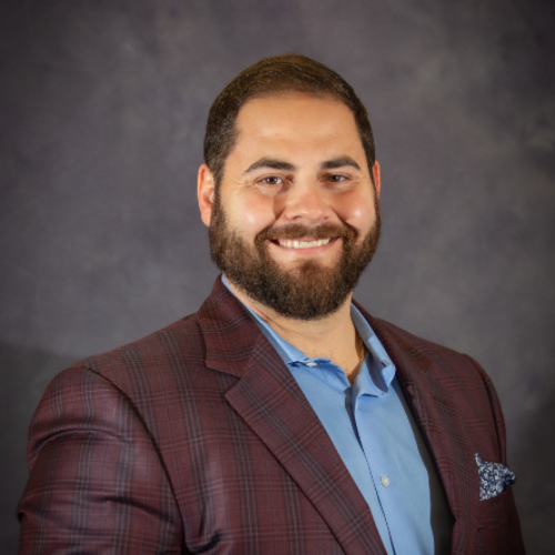 A man with a beard, in a blue shirt and maroon plaid blazer, smiles warmly—representing St. Paul's School on the Northshore.
