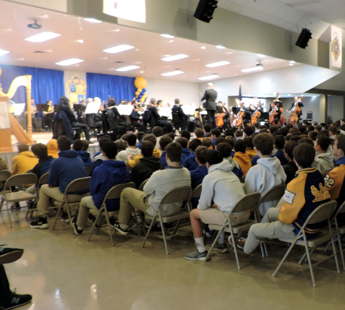 Students from St. Paul's School orchestra perform on stage as peers in uniforms watch attentively in the gymnasium.