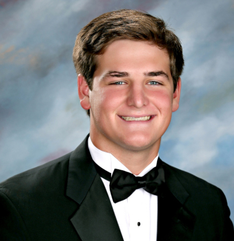 A young man in a black tuxedo and bow tie from St. Paul’s School smiles at the camera before a blue and gray studio backdrop.