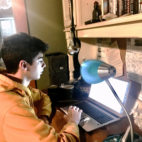 A student from St. Paul's School in a yellow hoodie works at a cluttered desk with a laptop, lamp, books, and papers in a dimly lit room.