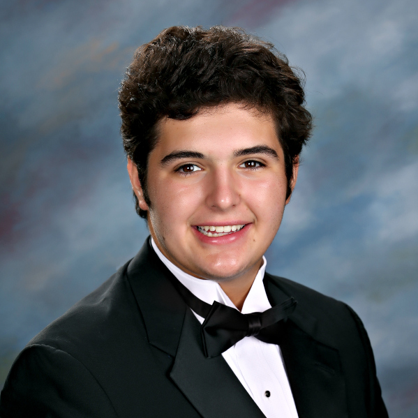 A young man with short, dark, curly hair in a black tuxedo smiles at the camera, representing St. Paul’s School against a blue studio background.