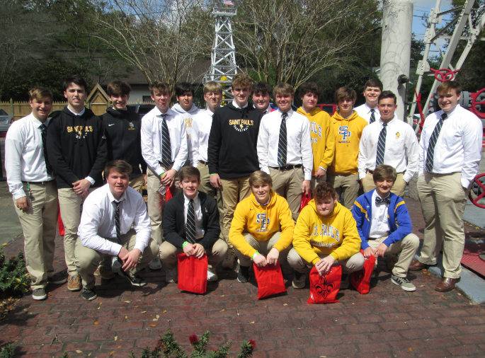 A group of 18 boys from St. Paul's School pose outdoors in school uniforms and sweatshirts, holding red gift bags on a brick walkway.