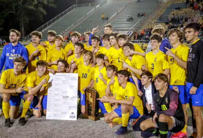 A boys' soccer team from St. Paul's School in yellow uniforms celebrates with a trophy and poster on the field, bleachers in the background.