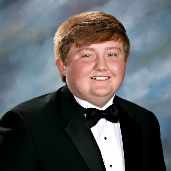 A young man with short light brown hair in a black tuxedo, posing in front of a blue-gray background at St. Paul’s School.