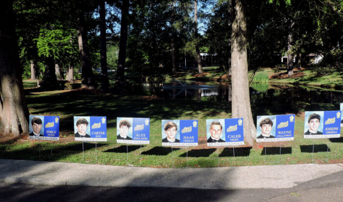 A row of graduation yard signs featuring St. Paul's School students lines a path in a park-like area with trees and a pond.