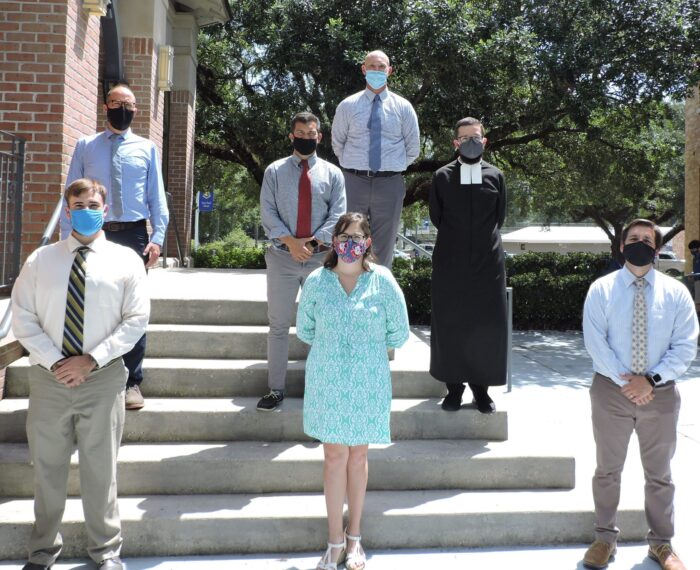Seven adults in masks pose on outdoor steps at St. Paul's School, a leading private school for boys on the Northshore, with trees and a building behind.