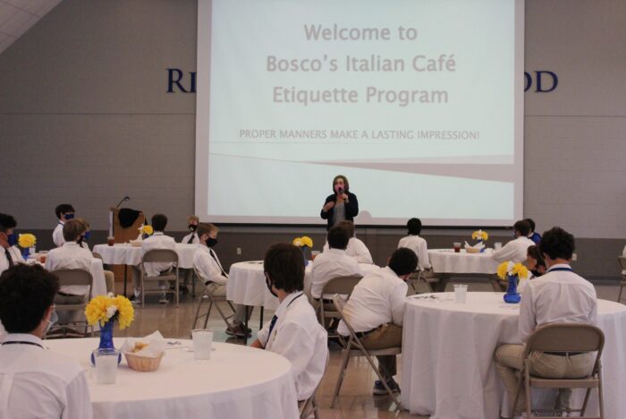 A woman speaks at a podium to students at round tables for Bosco’s Italian Café Etiquette Program at St. Paul’s School, displayed on screen.