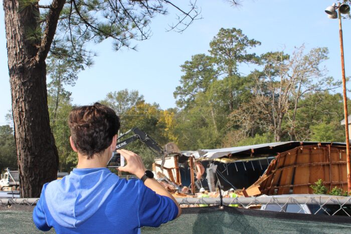 A person in a blue shirt takes a photo of a building near St. Paul's School being demolished by an excavator behind a fence.