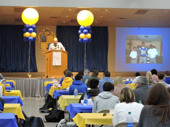 A man speaks at a podium on stage in a decorated hall at St. Paul's School, while guests watch a projected photo on the wall.