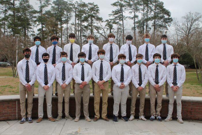 Eighteen young men from St. Paul's School in white shirts, ties, and masks stand outdoors before trees and a brick wall.