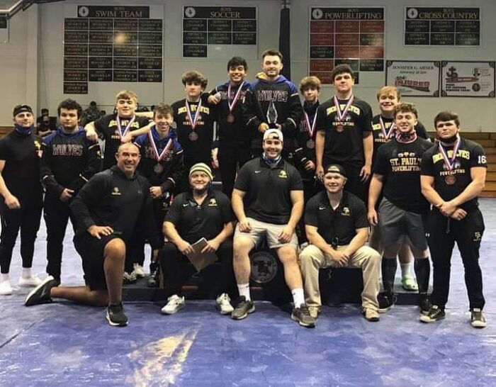 A group of St. Paul's School wrestlers and coaches pose indoors, some wearing medals, in front of banners at this school for boys on the Northshore.