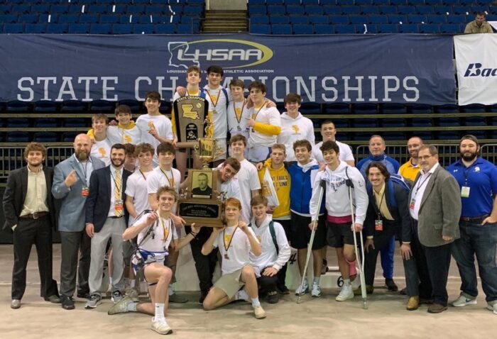 A boys’ team from St. Paul’s School poses indoors with a large trophy in front of an LHSAA State Championships banner, coaches, and staff.