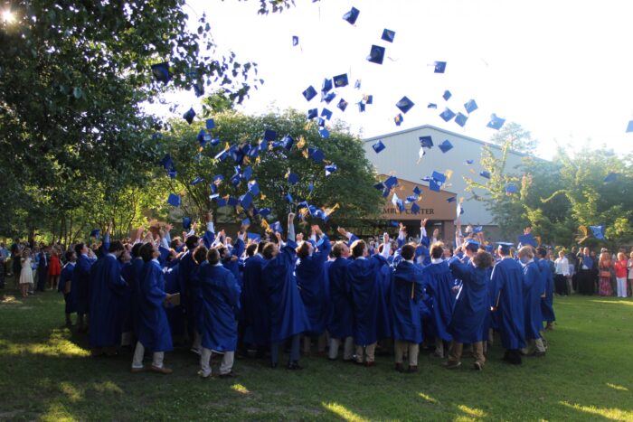 A group of graduates in blue gowns from St. Paul's School toss their caps in the air on a sunny day, surrounded by trees and people.