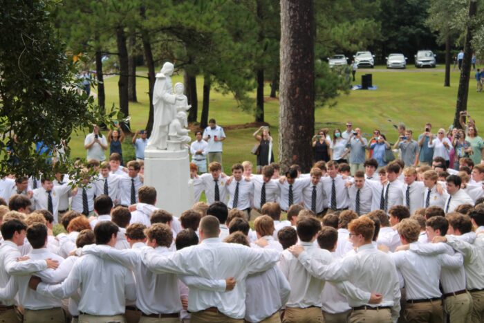 A large group of young men in white shirts and ties from St. Paul's School stand arm in arm around a statue, with photos being taken nearby.