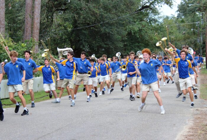 A marching band from St. Paul’s School in blue shirts and khaki shorts parades with instruments down a tree-lined street.