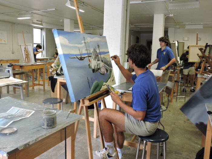 A student at St. Paul's School paints an airplane on canvas in an art classroom, with others working at easels in the background.
