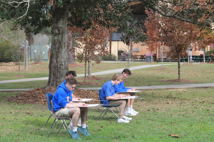 Four students in blue sweatshirts from St. Paul’s School sit outdoors, drawing or writing on pads in a grassy, tree-filled area.