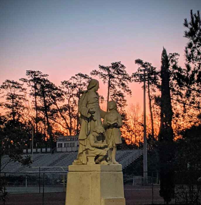 A stone statue of three people stands before empty bleachers at sunset on the campus of St. Paul’s School, northshore LA.