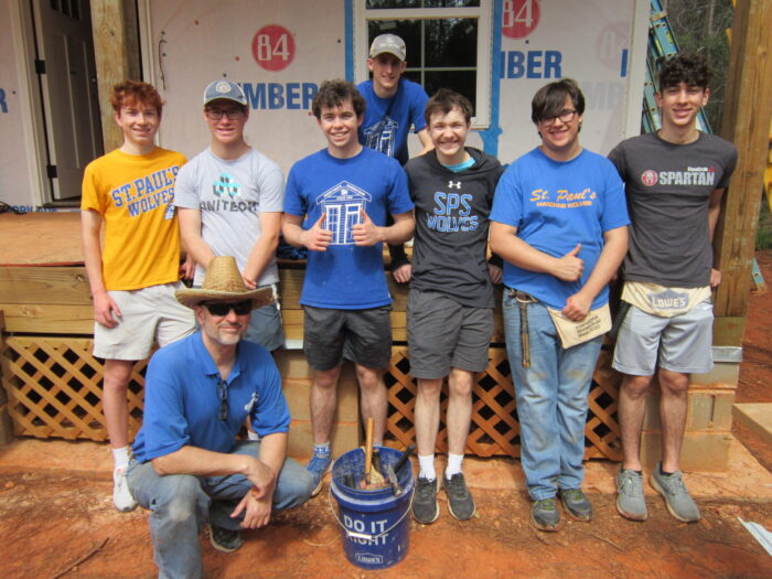 A group from St. Paul’s School poses in front of a house under construction; most are standing, one kneels, with a blue bucket in front.
