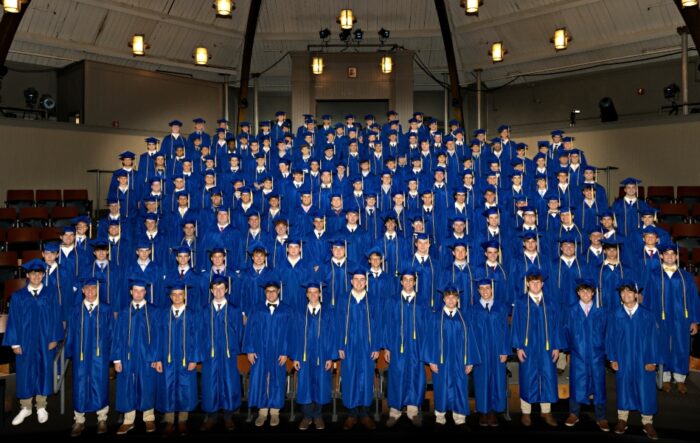 A large group of St. Paul's School graduates in blue caps and gowns stands in rows on a stage for a group photo in an auditorium.
