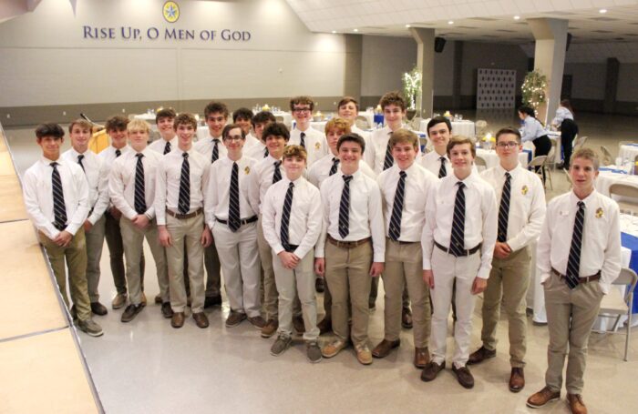 A group of young men from St. Paul's School in white shirts and ties stand smiling in a hall with a “RISE UP, O MEN OF GOD” sign.