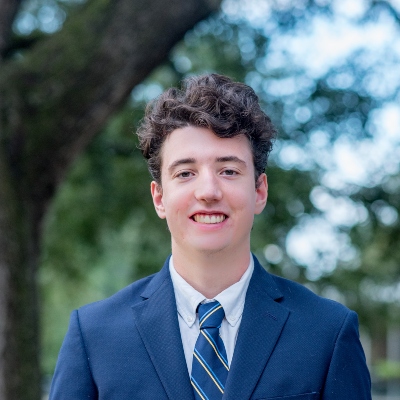 Young man with curly brown hair in a blue suit, dressed for a day at St. Paul's School, standing outdoors with trees in the background.