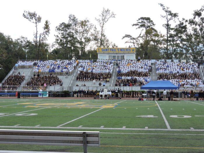 A large group gathers on bleachers at St. Paul’s School, a school for boys northshore LA, with tents set up on the field in front of them.