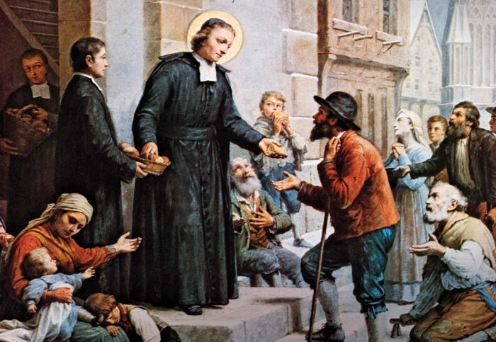 A man in religious attire hands bread to a begging man on crutches near St. Paul's School, surrounded by other needy people outside a building.