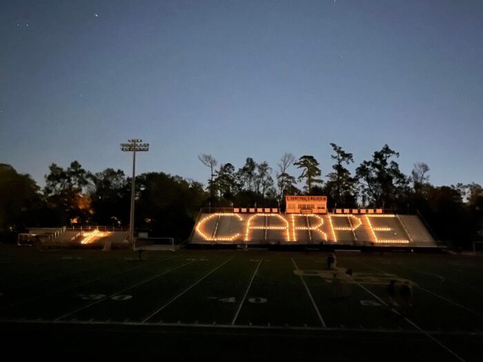 A stadium at dusk at St. Paul’s School, with bleachers lit to spell “CARPE”; trees and a scoreboard stand in the background.