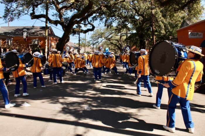 A marching band from St. Paul's School in yellow and blue uniforms plays instruments on a tree-lined street in the daytime.
