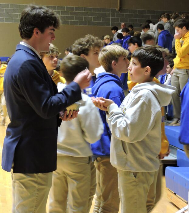 A group of boys in khaki pants and jackets talk in a gymnasium at St. Paul’s School near the blue bleachers.