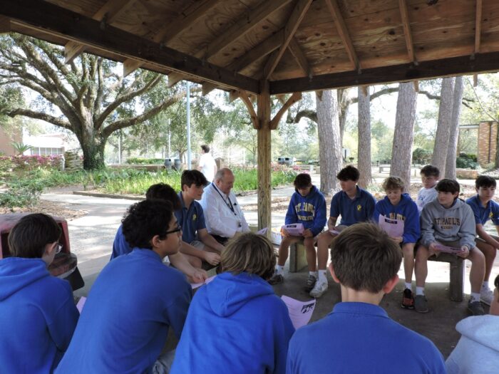 A group of boys and an adult from St. Paul's School sit in a circle under a wooden pavilion, holding papers, outdoors on a sunny day.