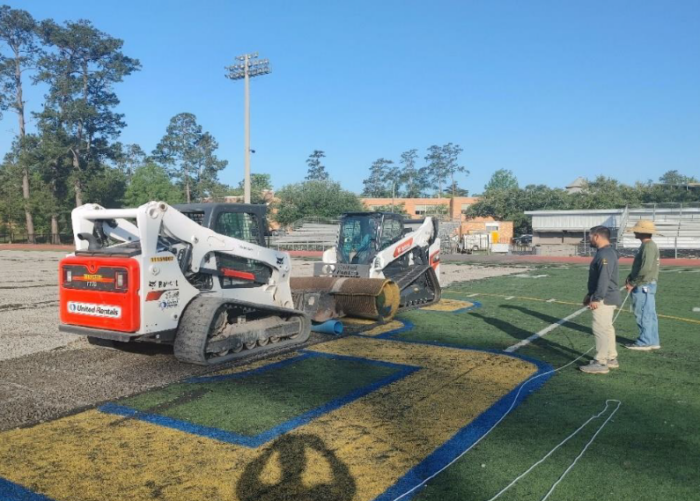 Two small construction vehicles work on the St. Paul's School sports field as three people stand nearby, holding ropes and observing.
