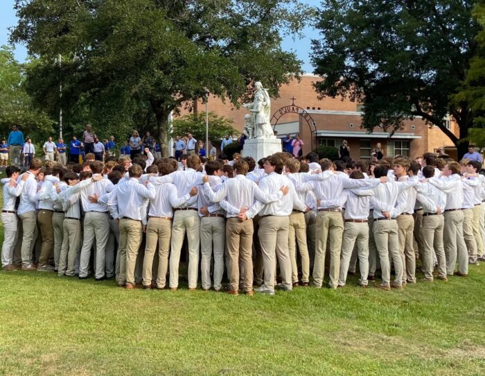 A large group of young men in khakis and white shirts stand arm-in-arm near a statue at St. Paul’s School, a school for boys northshore LA.