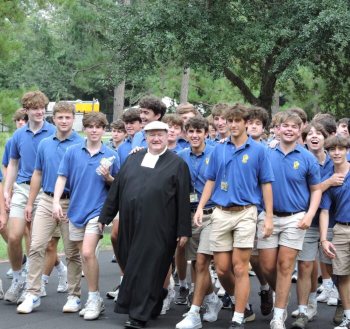 A group of teenage boys from St. Paul’s School walk outside in blue polos and khakis with a man in a black robe and white collar at the center.
