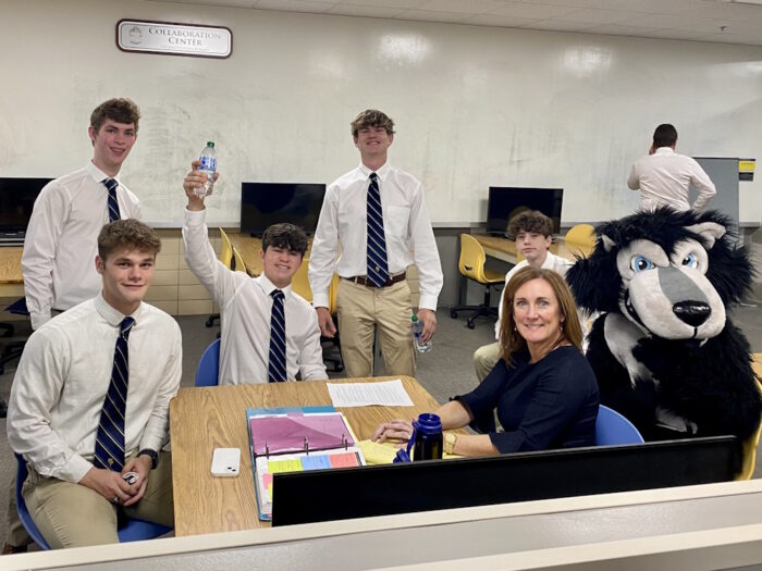 Six students from St. Paul's School, a school for boys on the Northshore, sit with a woman and a mascot in a room with computers and a whiteboard.