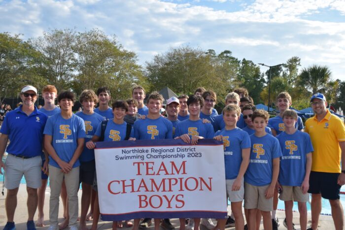 A St. Paul’s School boys’ swim team poses outdoors with a “TEAM CHAMPION BOYS” banner at a swimming championship event.