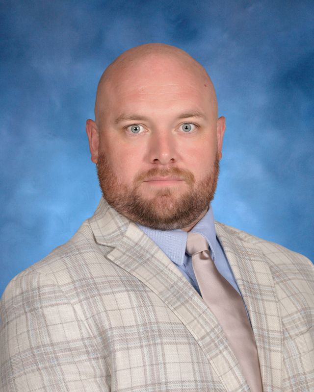 A man with a bald head and beard in a plaid suit jacket, blue shirt, and beige tie poses for St. Paul’s School, a school for boys northshore LA.