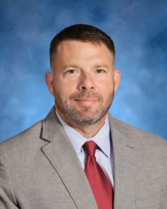 A man with short brown hair and a beard, in a gray suit and red tie, poses before a blue background at St. Paul’s School.