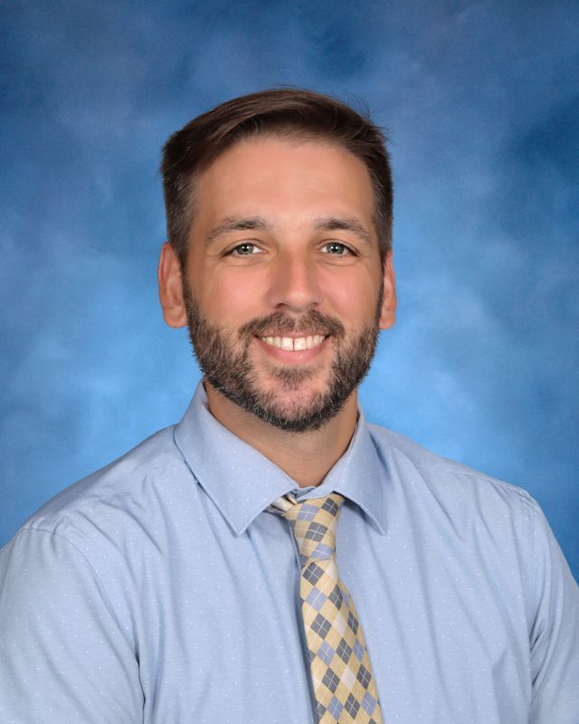 A man with short brown hair and a beard, in a light blue shirt and tie, poses before a blue background at St. Paul’s School.