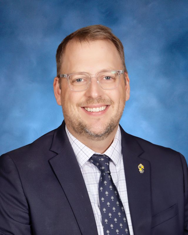 A man in a suit, glasses, and tie is smiling in front of a blue studio background at St. Paul’s School.