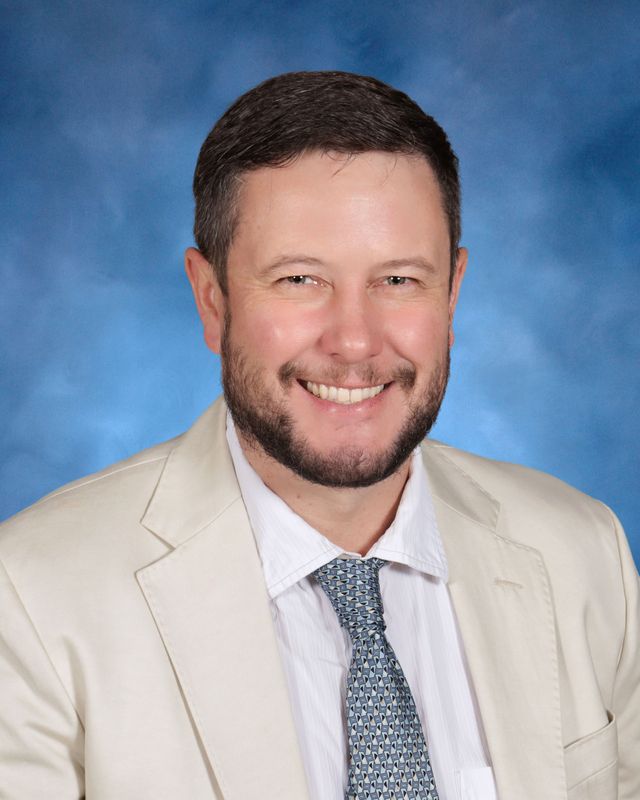 A man with short dark hair and a beard smiles, dressed for St. Paul's School, a top school for boys northshore LA, against a blue background.