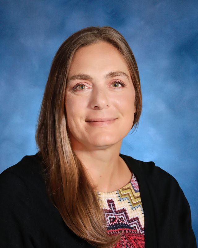 A woman with long brown hair smiles at the camera, wearing a patterned top and black cardigan, with a blue St. Paul's School background.