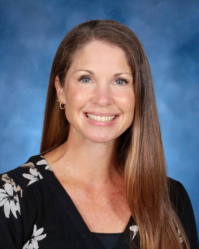 Woman with long straight brown hair, wearing a black floral top, smiling in front of a blue background at St. Paul's School.
