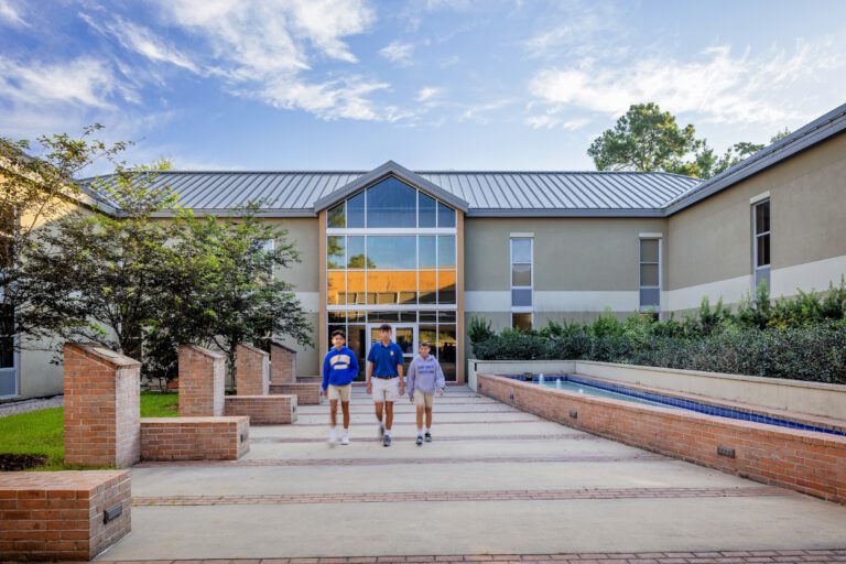 Three people walk toward St. Paul's School, a private school on the Northshore, past brick columns and modern windows under a blue sky.