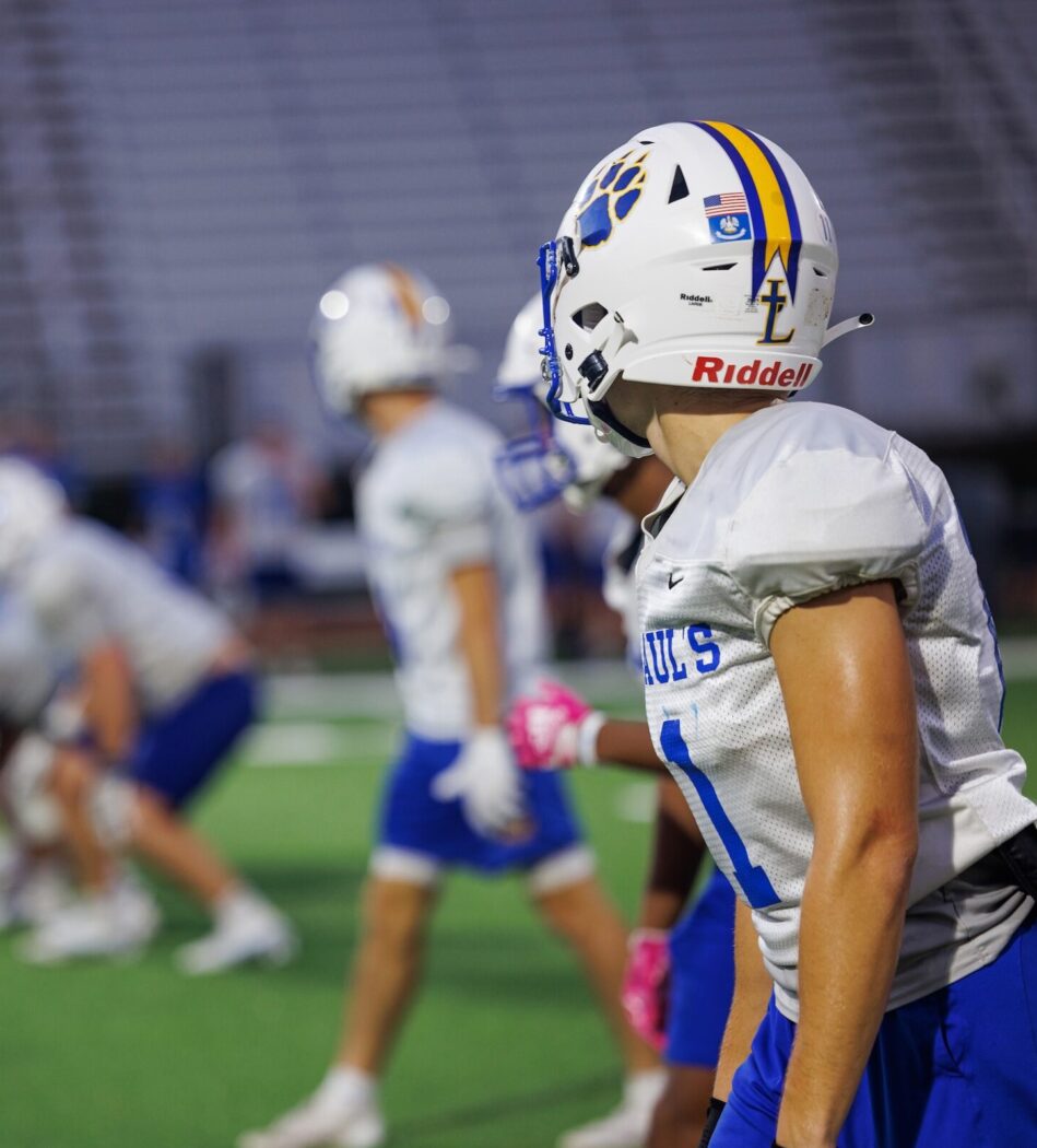 A group of St. Paul's School football players practice on a field; one player in focus stands to the right, watching the action.