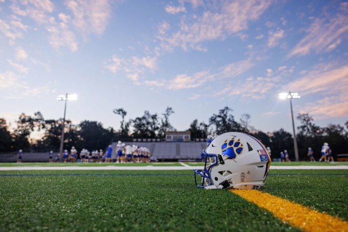 A white St. Paul's School football helmet with a blue paw print sits on green turf, players and stadium lights in the background.